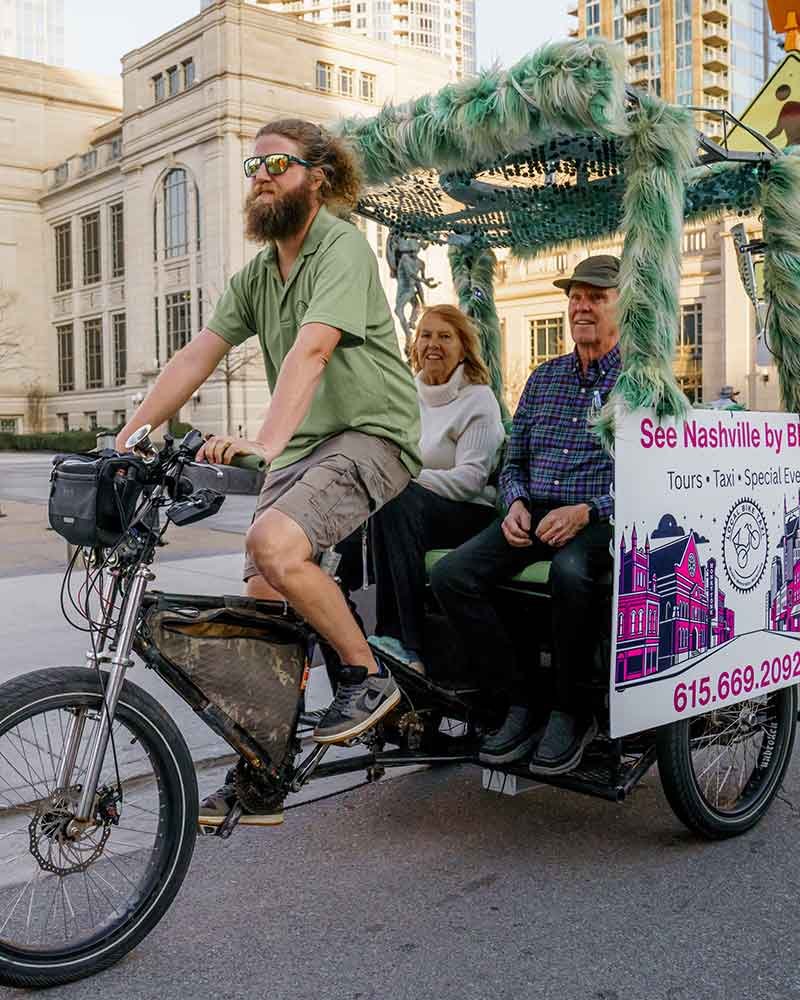 An elderly couple riding on a green furry neon pedicab with a hippie looking guy pedaling the bike.