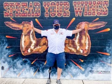Man posing in front of a mural of giant chicken wings. The wings are set up to look like angel chicken wings.