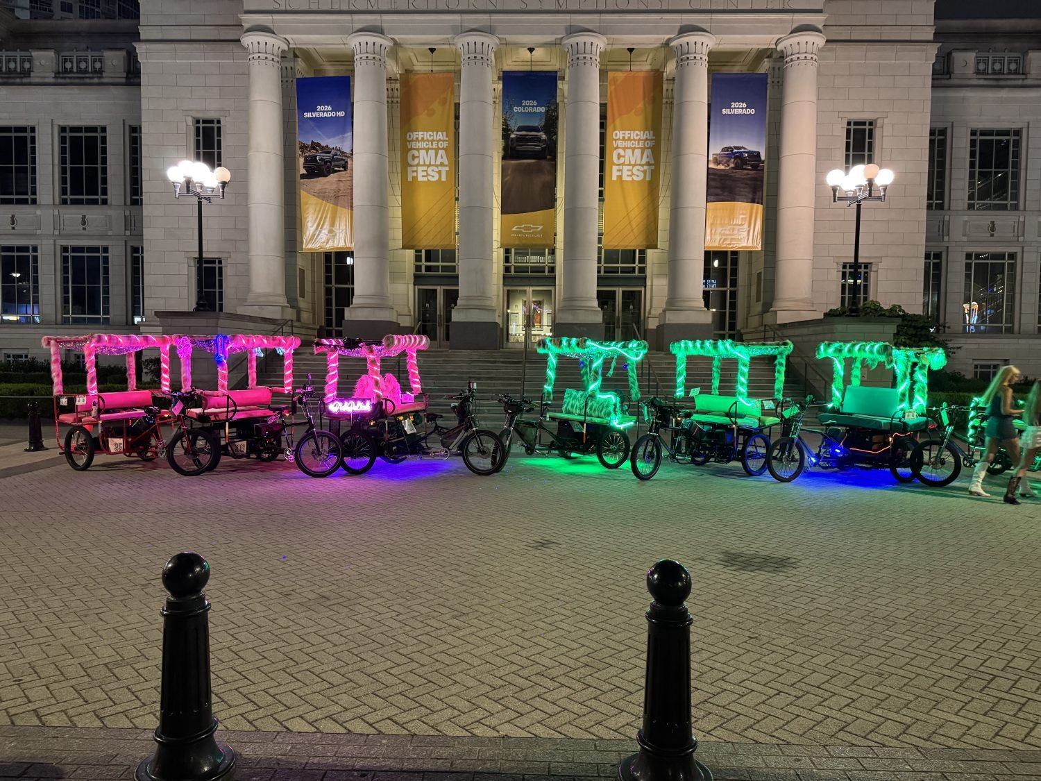 A picture of pink and green furry 6 seating bill board pedicabs in Nashville, in front of some CMA fest banners.