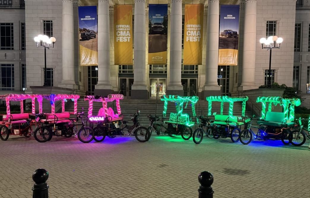 A picture of pink and green furry 6 seating bill board pedicabs in Nashville, in front of some CMA fest banners.