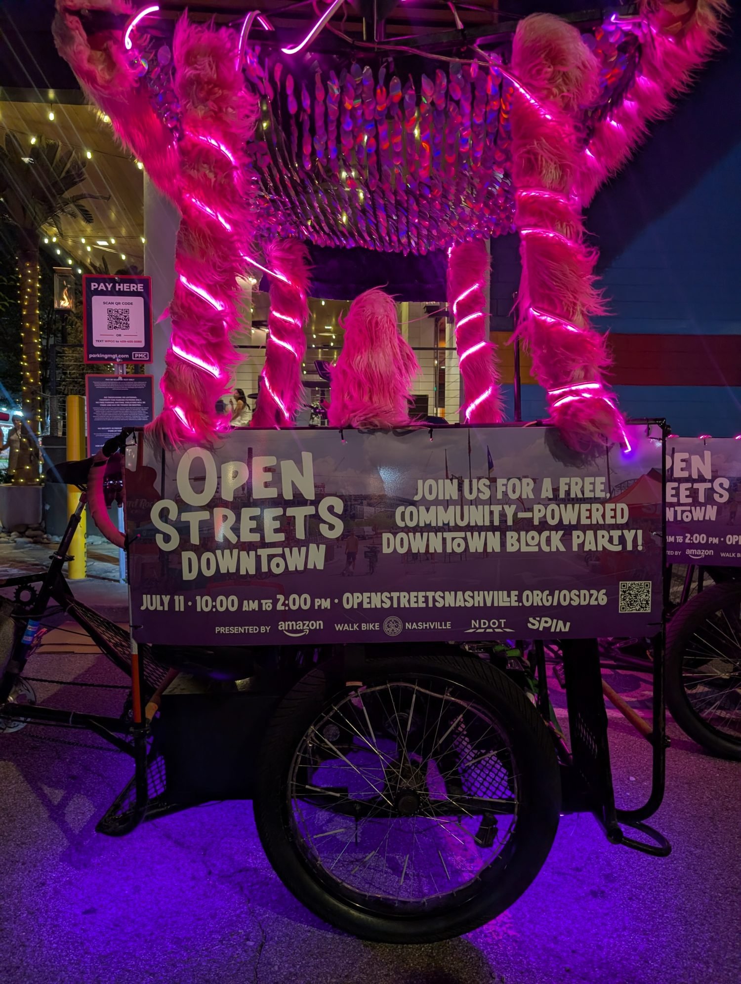 A pink pedicab that seats 6 people. It has one bench facing forward and another facing rear. The passengers sit back to back. This one has and advertisement on the side, and pink neon lights