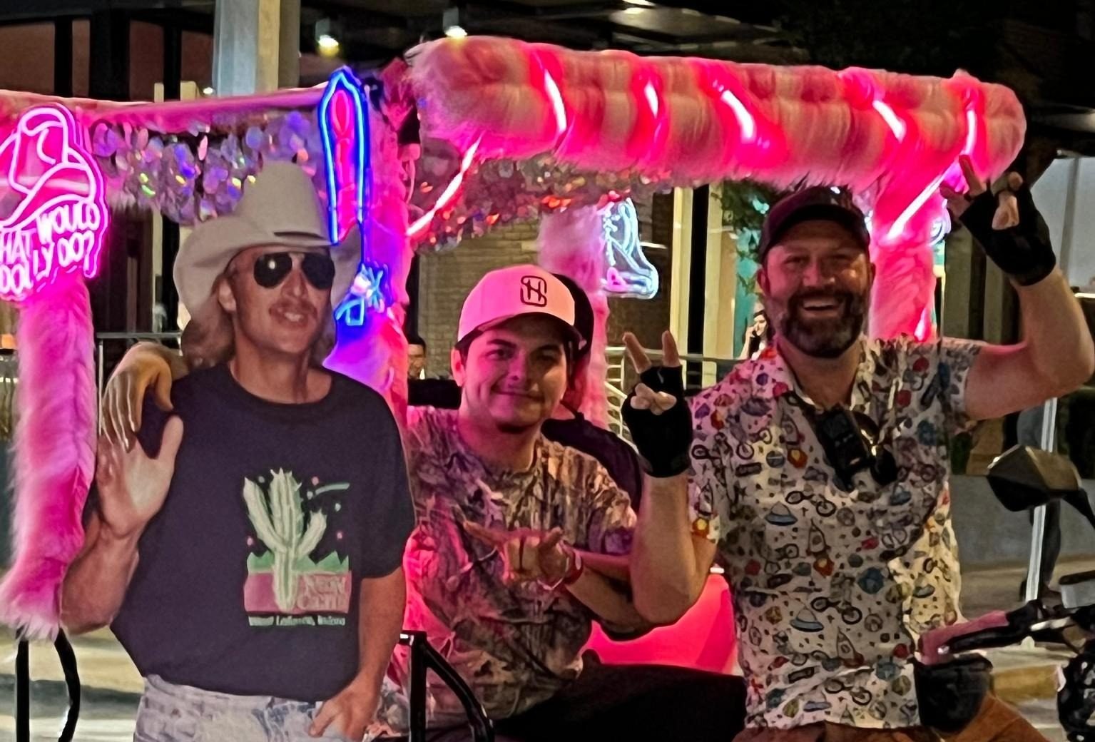 Guys riding on a hot pink neon pedicab with pink fur with giant life sized Alan Jackson Cut out.