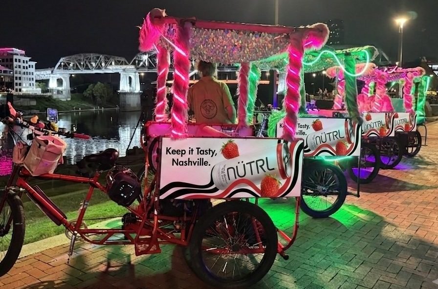 Pedicabs that are pink and green neon with pink and green furry shiny canopies, on the river front in Nashville, TN with NUTRL beverage ads on them.